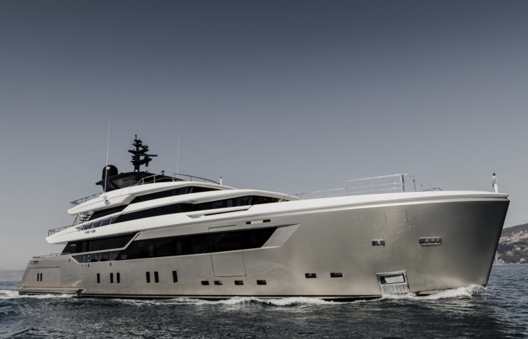 A sleek, modern luxury yacht with a silver hull cruises on calm blue waters under a clear sky, with distant land visible in the background.
