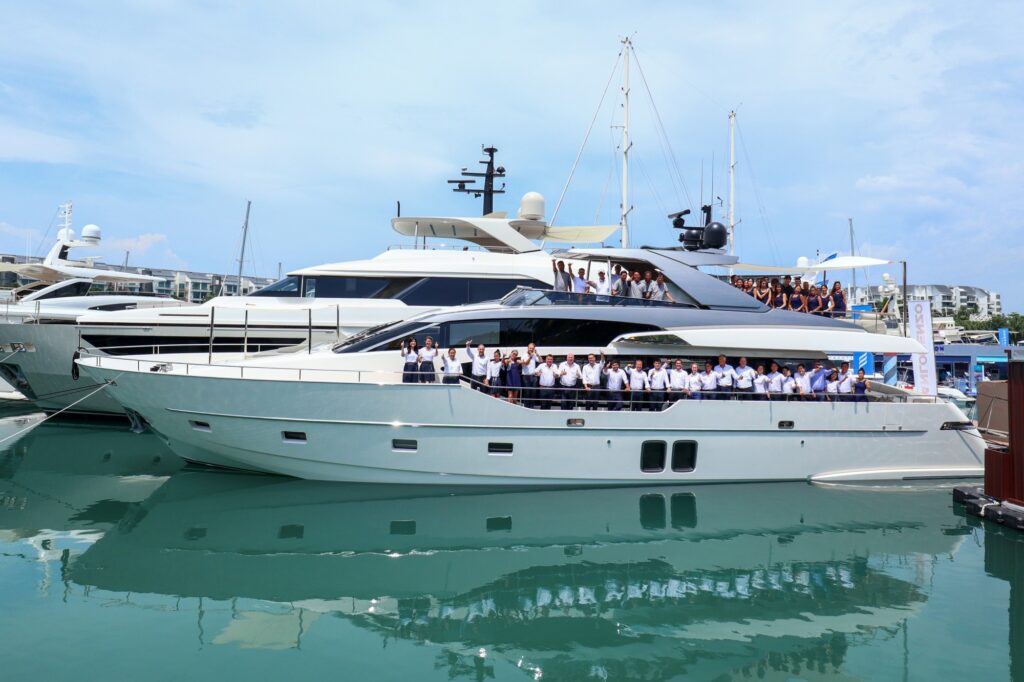 A large group of people dressed in white uniforms stand on a docked luxury yacht, with another yacht and marina buildings visible in the background under a partly cloudy sky.