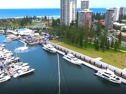 A marina with several moored yachts, bordered by a grassy park and tall modern buildings, with the sea visible in the background on a clear day.
