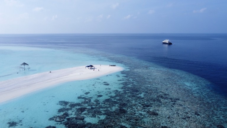 A small sandy island with a few people and structures sits amid clear blue water and coral reefs, with a yacht anchored nearby and a vast expanse of ocean under a blue sky.