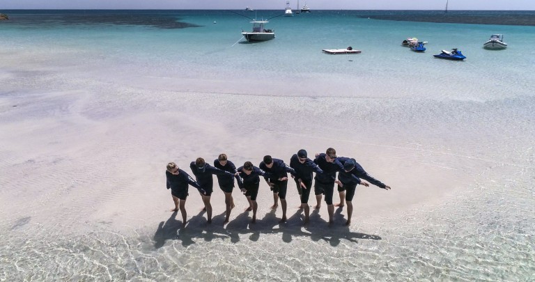 A group of eight people stands in shallow, clear water at the beach, holding hands in a line. Several boats and jet skis float in the calm turquoise sea in the background under a clear sky.