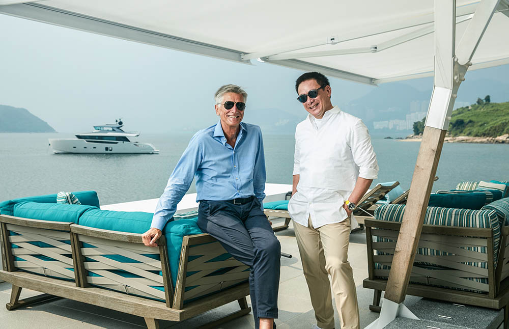 Two men in sunglasses are smiling and posing on a waterfront patio with turquoise seating; a yacht is in the water and hills are visible in the background under a hazy sky.