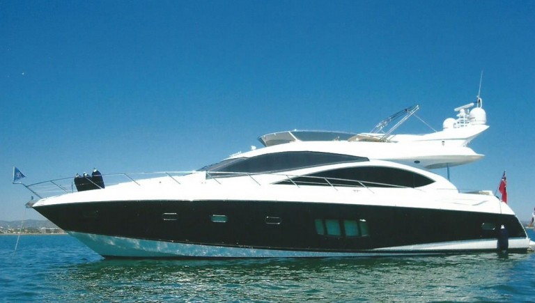 A sleek, modern yacht with a white upper deck and dark hull floats on calm blue water under a clear sky. The yacht has several windows, and flags are visible at the stern.