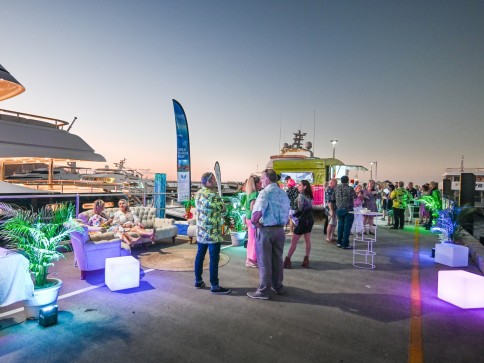 People socialising at a dockside event during sunset, with yachts in the background, illuminated plants and furniture, and a relaxed, festive atmosphere.