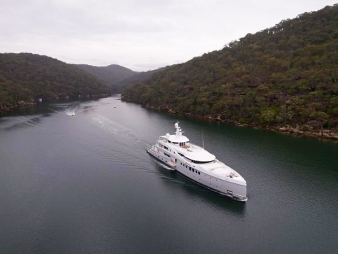 A large white yacht cruises along a narrow, calm river surrounded by lush, green hills under an overcast sky. Another smaller boat is visible in the distance.