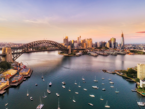 Aerial view of Sydney Harbour at sunset, showing the Sydney Harbour Bridge, city skyline, and boats moored in the water with soft pink and orange hues in the sky.