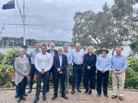 A group of eleven people, dressed in business and casual attire, stand outdoors on a brick patio with greenery, a flagpole, and a body of water with boats in the background under a cloudy sky.