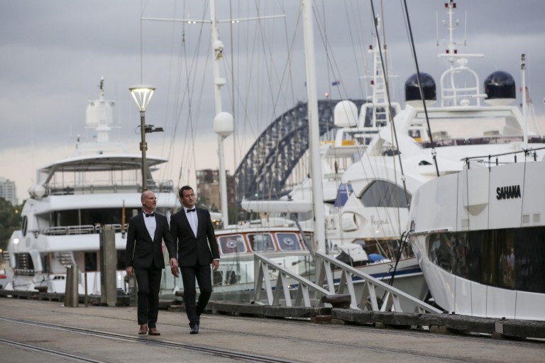 Two men in tuxedos walk along a quay lined with luxury yachts, with a cloudy sky and the metal arch of a large bridge visible in the background.