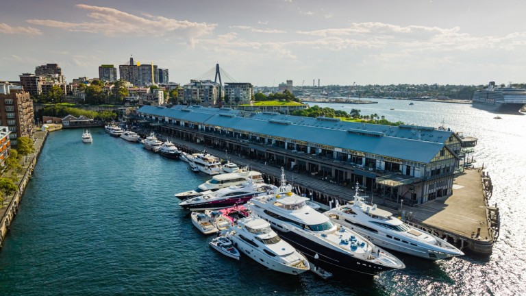 Several luxury yachts are moored at a long quay beside a city waterfront under a partly cloudy sky, with buildings and a bridge visible in the background.