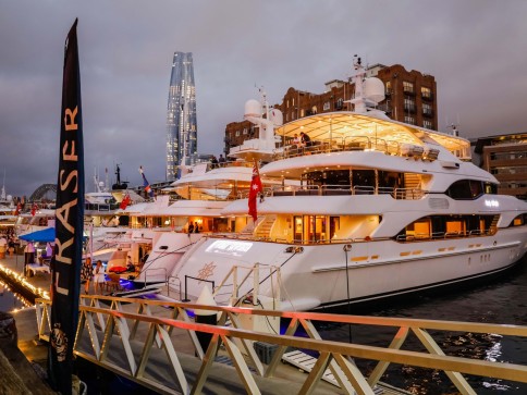 Luxury yachts moored at a marina during dusk, with city buildings and a modern glass skyscraper in the background. Warm lights illuminate the boats, creating a vibrant atmosphere under a cloudy sky.