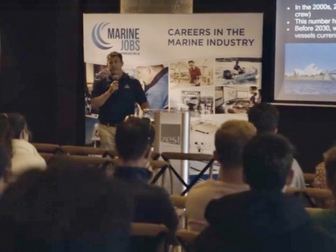 A speaker presents to an audience at a seminar about careers in the marine industry, with banners and a projection screen displaying information in the background.
