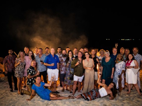 A group of about 30 adults, dressed in summer attire, pose and smile together on a sandy beach at night. Some hold drinks, and a bonfire glows behind them under a dark sky.