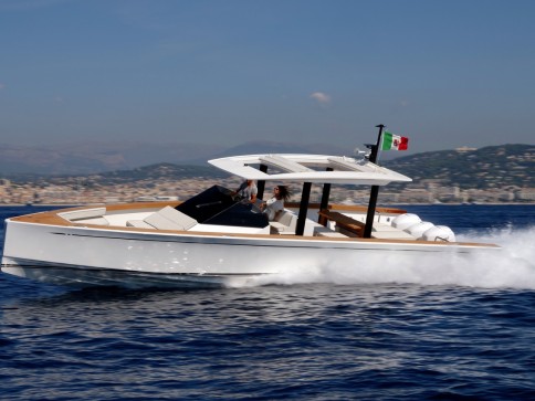 A sleek white motor yacht speeds across the blue water, creating a trail of splashes. Two people are seated at the helm, and an Italian flag flies at the stern with a coastal city and mountains visible in the background.