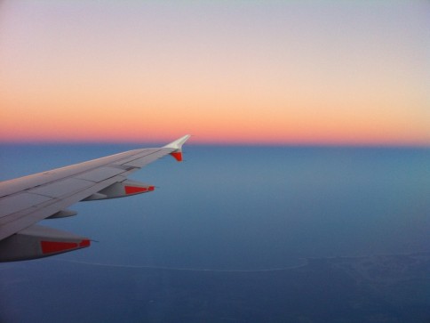 View from an aeroplane window showing the wing against a colourful sky at sunset, with shades of pink, orange, and blue blending above a distant coastline.