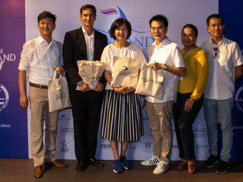 Six people stand together smiling and holding canvas tote bags at an event, posing in front of a blue and white backdrop with various logos and text.