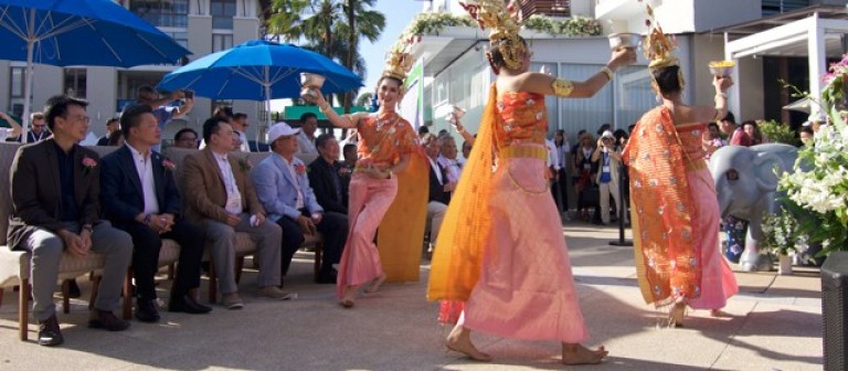 Three women in traditional Thai attire perform a dance outdoors for an audience of seated men in suits. Blue umbrellas provide shade, and a decorated elephant statue is visible on the right.