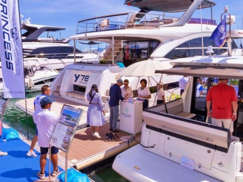 People gather on a quay next to luxury yachts at a boat show. Some are talking to staff under a parasol, while others look at the boats. Promotional banners and sunny weather are visible.