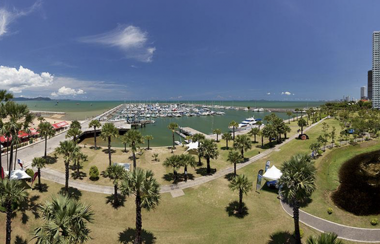 A marina filled with boats alongside a green park with palm trees, walking paths, and tents under a clear blue sky. High-rise buildings are visible on the right side of the image.