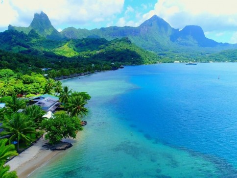 Aerial view of a tropical coastline with turquoise water, lush green mountains, palm trees, and small houses near the beach under a partly cloudy sky.