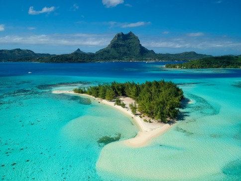 A small, lush island with white sandy beaches sits in clear turquoise water, with a larger island and a tall mountain visible in the background under a bright blue sky.