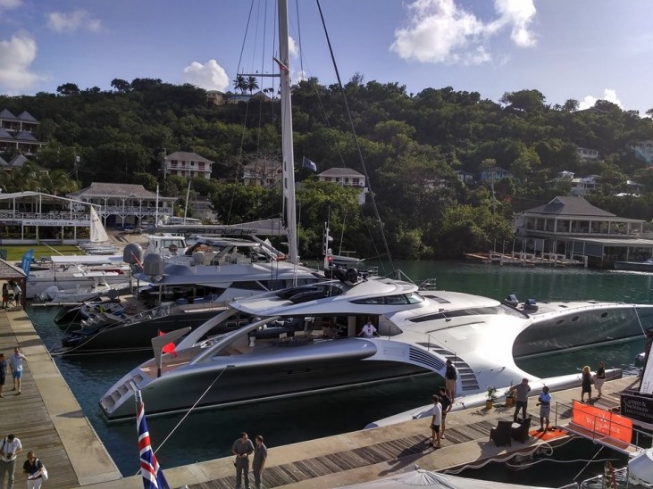 Several luxury yachts, including a large, futuristic silver trimaran, are moored at a marina with people walking on the pontoons. Lush green hills and elegant houses are visible in the background under a partly cloudy sky.