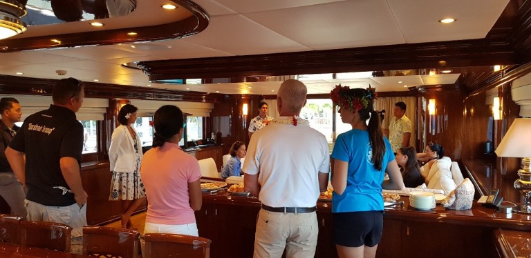 A group of people are gathered inside a luxurious yacht, talking and socialising around a table with food. Some are standing, others are seated, and the yacht’s polished wood interior is visible.