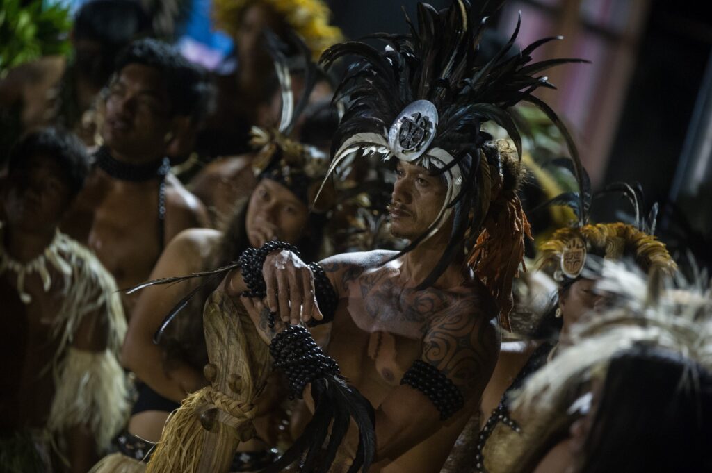 A group of people in traditional Polynesian attire with feathered headdresses and body ornaments stand together, some with tattoos, participating in a cultural event or ceremony.