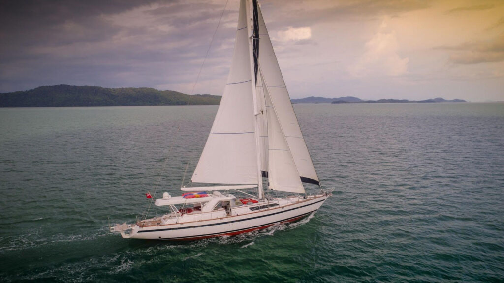 A white sailing boat with raised sails glides across calm, open water near a distant, tree-covered shoreline under a cloudy sky with a hint of sunset light.