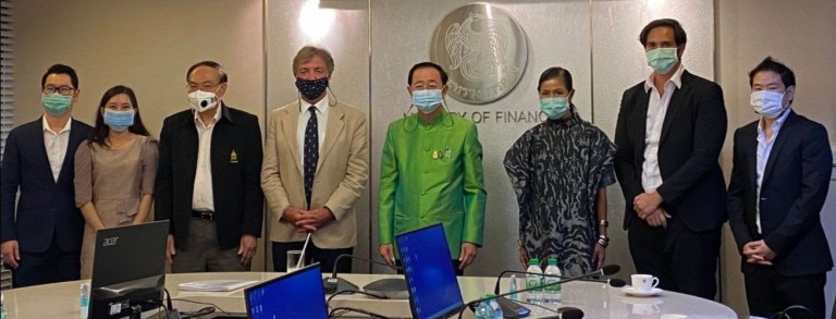 Eight people wearing face masks stand in a conference room in front of a wall with a Ministry of Finance logo. Laptops and water bottles are on the table in front of them.