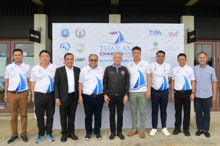 Nine men stand in a row in front of a Thailand Charter Week banner, wearing coordinated white and blue shirts with sailing logos, posing for a group photo outside a building.