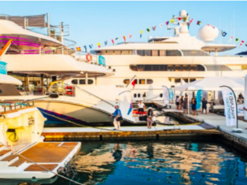 Several large white yachts are moored at a marina, with people walking on the pier between them. Colourful flags are strung above one yacht, and the water reflects the boats and bright sky.