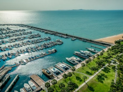 Aerial view of a marina filled with numerous yachts and boats moored in clear blue water, adjacent to a green park with rows of palm trees and a sandy beach along the sea.
