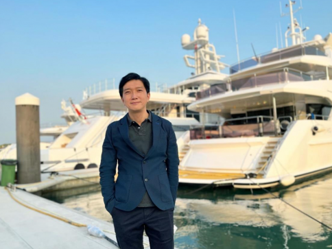 A man in a navy blazer and dark trousers stands smiling on a marina quay, with several large white yachts moored behind him under a clear blue sky.