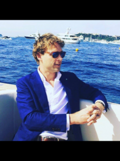 A man wearing sunglasses, a white shirt, and a blue blazer sits relaxed on a boat with yachts and coastline visible in the background on a sunny day.