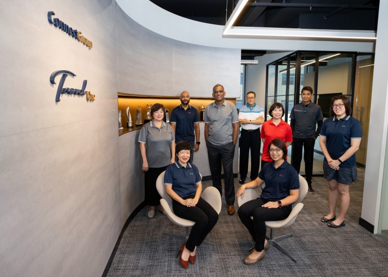 Nine people, a mix of men and women, pose for a group photo in a modern office foyer. Two women are seated, while the rest stand behind them near a trophy display and company signage on the wall.