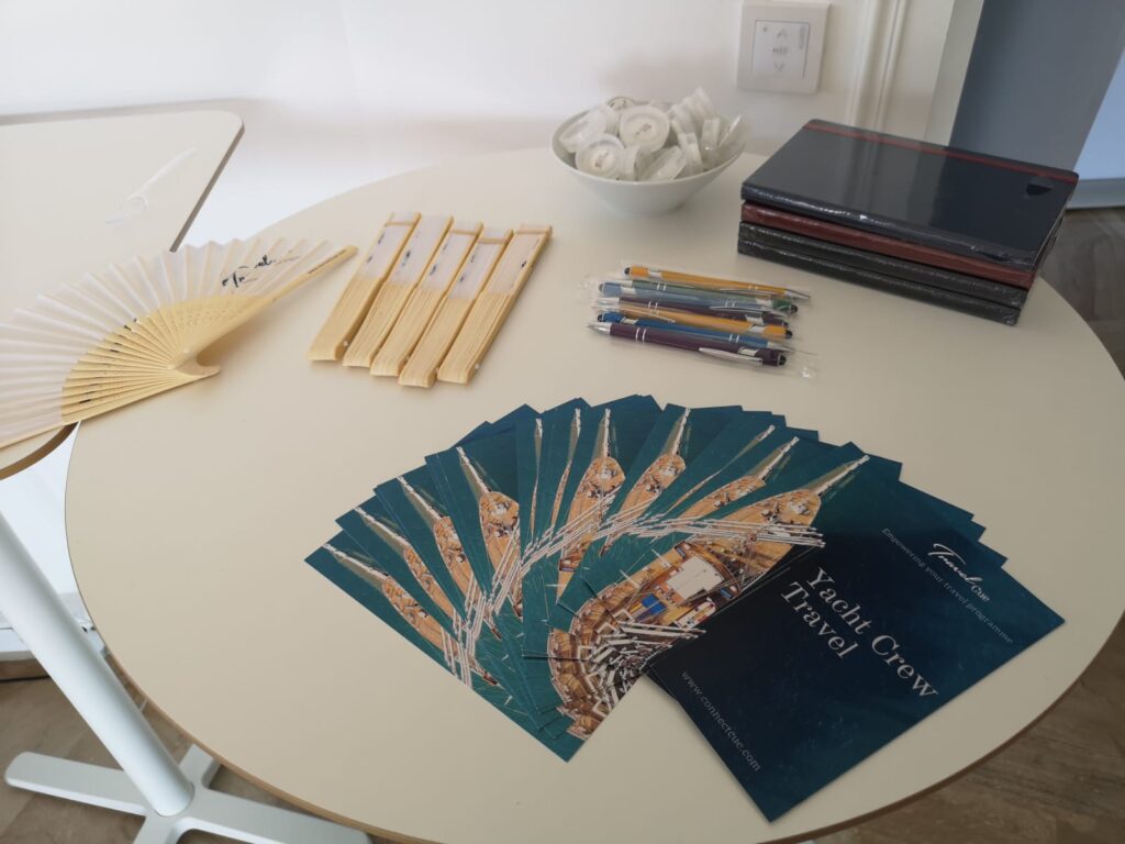 A round table displaying brochures titled Yacht Crew Travel, several pens, unfolded and folded handheld fans, a stack of folders, and a bowl of individually wrapped round items.