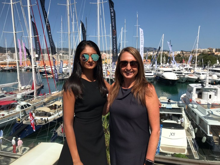 Two women wearing sunglasses and sleeveless dark dresses stand smiling in front of a marina filled with boats and yachts, with masts and flags visible behind them under a clear blue sky.