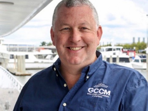 A smiling man in a blue button-up shirt that reads GCCM stands outdoors near a marina with several boats and yachts in the background.