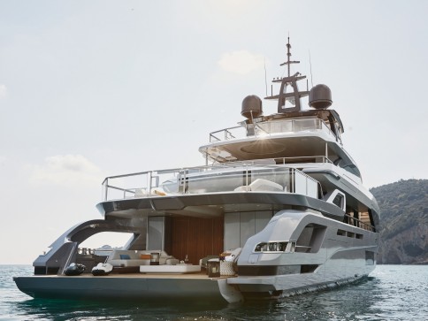 A luxury yacht is anchored on calm water near a tree-covered coastline, seen from the rear and featuring multiple decks with outdoor seating and sunbathing areas under a clear sky.