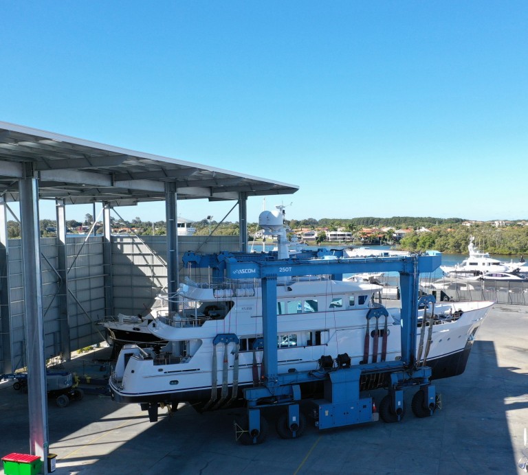 A large white yacht is being transported by a blue boat lift in a marina facility, partially sheltered by a tall metal roof structure, with trees and other boats visible in the background under a clear blue sky.