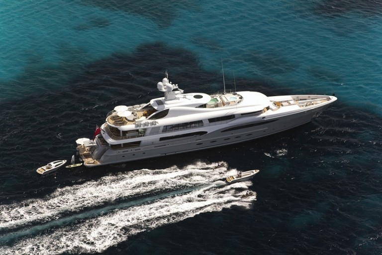 Aerial view of a large white luxury yacht anchored in clear blue water, with two smaller speedboats creating white wake trails as they move near the yacht.
