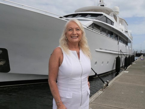 A woman with long blonde hair, wearing a sleeveless white dress, stands on a quay in front of a large white yacht on a cloudy day.