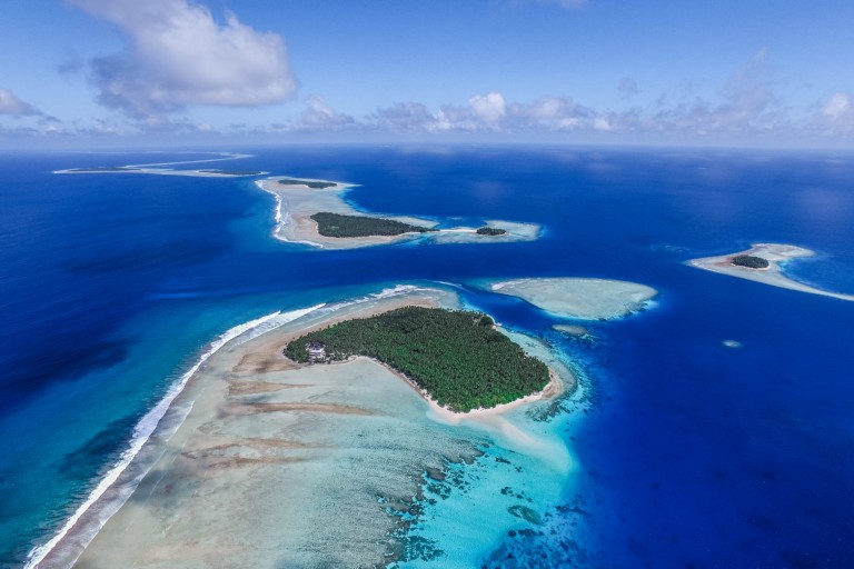 Aerial view of small tropical islands surrounded by turquoise water, coral reefs, and white sandy beaches, with lush green vegetation and clear blue sky above.