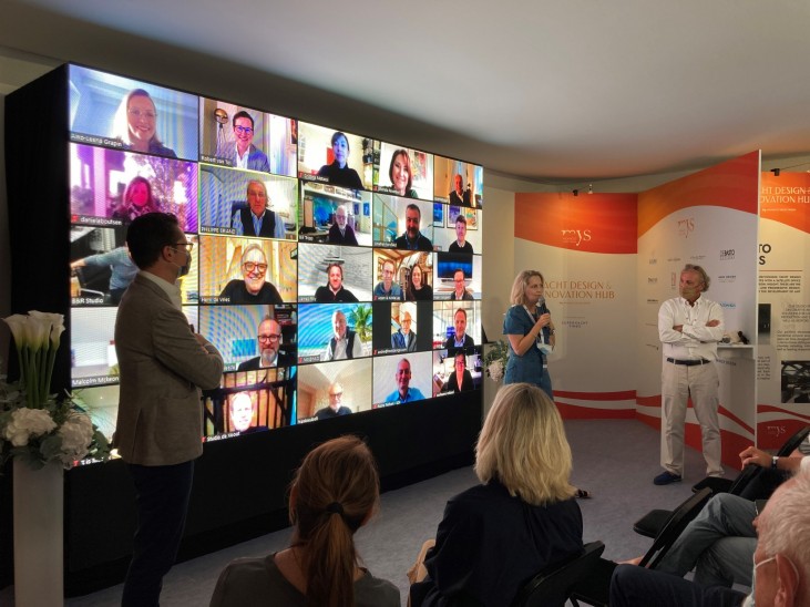 People seated in a room watch a large video wall displaying many participants on a video call; three individuals stand in front of the screen, two speaking whilst one observes. Exhibition panels are visible in the background.
