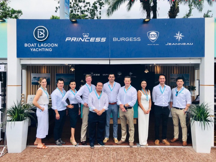 A group of eleven people, dressed in white and light-coloured clothing with blue lanyards, stand smiling in front of a stand displaying yacht company logos at an outdoor event.