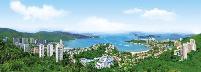 Panoramic view of a coastal city with tall buildings surrounded by green hills, curving shoreline, blue water, and distant islands under a bright, partly cloudy sky.