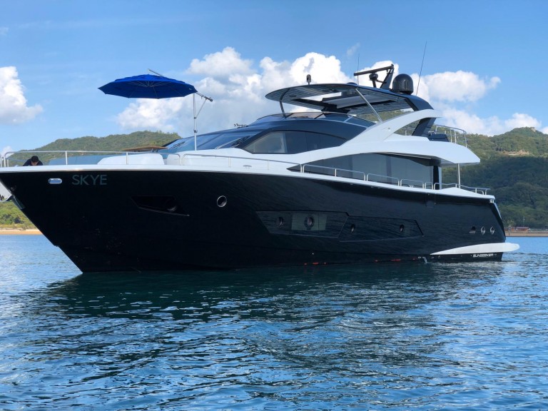 A sleek black and white luxury yacht named SKYE floats on calm blue water, with a blue umbrella on the upper deck. Hills and a partly cloudy sky are visible in the background.
