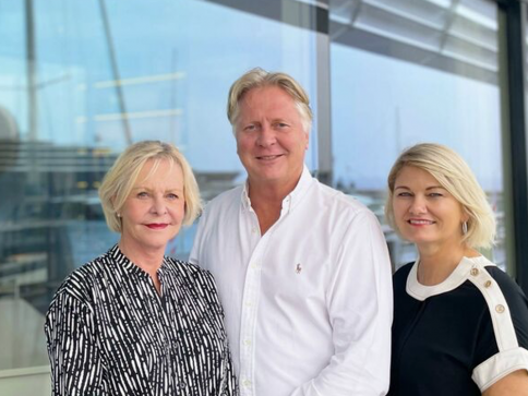 Three adults stand closely together and smile at the camera outside by a glass wall, with a marina and boats visible in the background. All are wearing casual, light-coloured clothing.