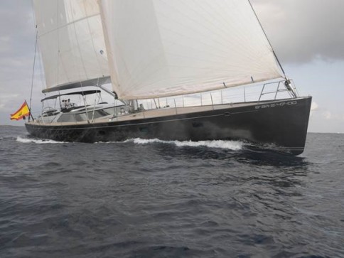 A sleek, modern sailboat with white sails moves through the sea, flying a Spanish flag at the stern under a cloudy sky.