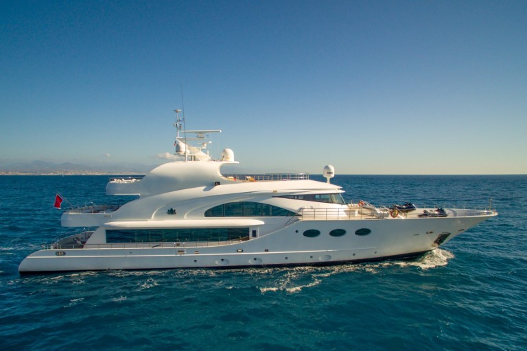 A large white luxury yacht sails on calm blue ocean water under a clear sky, with land faintly visible in the distant background.